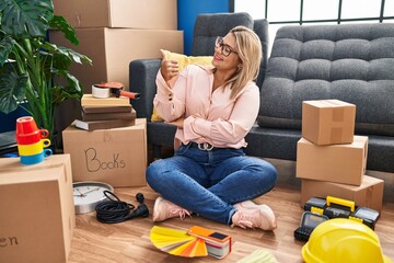 Wall Mural - Young hispanic woman moving to a new home sitting on the floor looking proud, smiling doing thumbs up gesture to the side