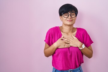 Poster - Young asian woman with short hair standing over pink background smiling with hands on chest with closed eyes and grateful gesture on face. health concept.