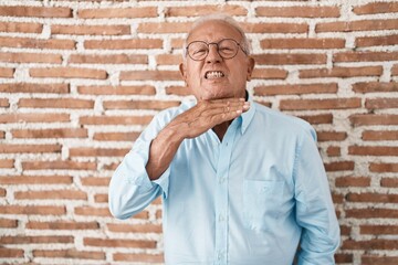 Poster - Senior man with grey hair standing over bricks wall cutting throat with hand as knife, threaten aggression with furious violence