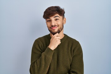 Canvas Print - Arab man with beard standing over blue background looking confident at the camera smiling with crossed arms and hand raised on chin. thinking positive.