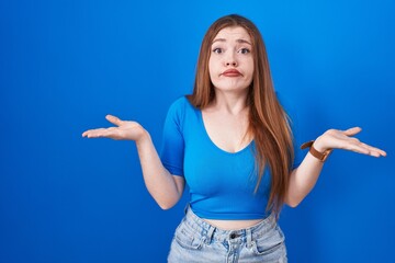 Canvas Print - Redhead woman standing over blue background clueless and confused expression with arms and hands raised. doubt concept.