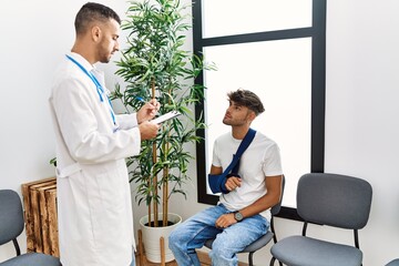 Wall Mural - Two hispanic men doctor and patient writing on clipboard sitting on chair at hospital waiting room