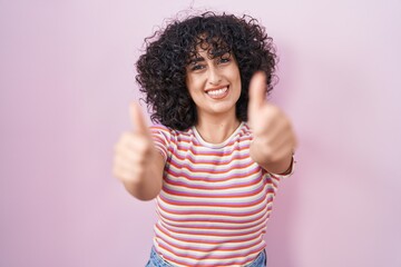 Poster - Young middle east woman standing over pink background approving doing positive gesture with hand, thumbs up smiling and happy for success. winner gesture.