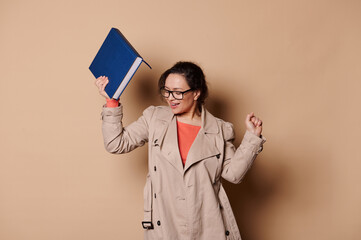 Cheerful joyful middle-aged woman, wearing beige coat and stylish spectacles, teacher educator celebrates the school year's end, dancing with a hardcover book in her hand, on isolated beige background