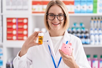 Canvas Print - Young caucasian woman working at pharmacy drugstore holding pills an piggy bank smiling with a happy and cool smile on face. showing teeth.