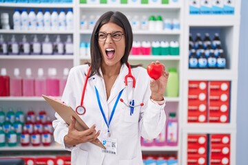 Poster - Brunette young woman working at pharmacy drugstore holding red heart angry and mad screaming frustrated and furious, shouting with anger. rage and aggressive concept.