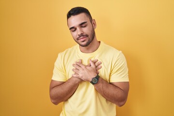 Canvas Print - Young hispanic man standing over yellow background smiling with hands on chest with closed eyes and grateful gesture on face. health concept.