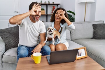 Wall Mural - Hispanic middle age couple sitting on the sofa using computer laptop smiling happy doing ok sign with hand on eye looking through fingers