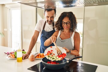 Wall Mural - Middle age hispanic couple smiling confident pouring food on frying pan at kitchen