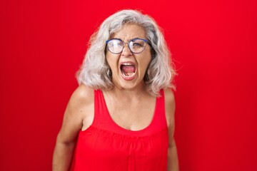 Poster - Middle age woman with grey hair standing over red background angry and mad screaming frustrated and furious, shouting with anger. rage and aggressive concept.