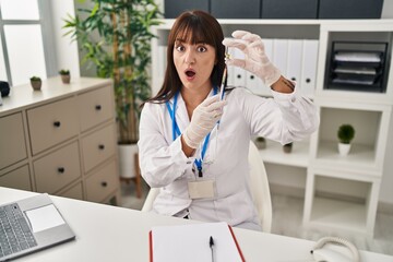 Sticker - Young brunette doctor woman holding syringe and vaccine afraid and shocked with surprise and amazed expression, fear and excited face.