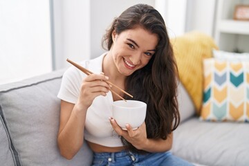 Young hispanic woman eating chinese food sitting on sofa at home