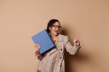 Joyful middle-aged pretty woman educator, school teacher in stylish beige coat and spectacles, celebrating the end of academic year, dancing with a hardcover book in hand, isolated cream background