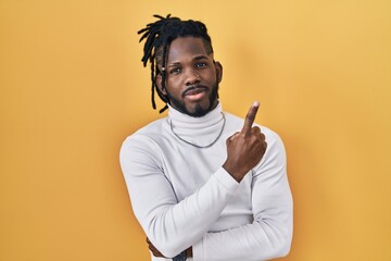 Sticker - African man with dreadlocks wearing turtleneck sweater over yellow background pointing with hand finger to the side showing advertisement, serious and calm face