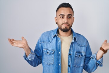 Sticker - Young hispanic man standing over isolated background clueless and confused expression with arms and hands raised. doubt concept.