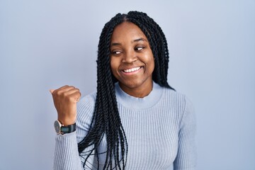 Wall Mural - African american woman standing over blue background smiling with happy face looking and pointing to the side with thumb up.