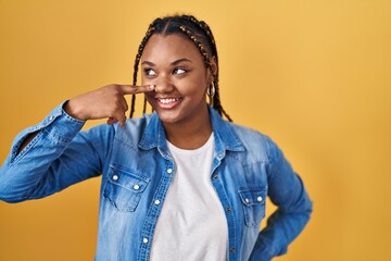 Poster - African american woman with braids standing over yellow background pointing with hand finger to face and nose, smiling cheerful. beauty concept