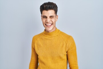 Wall Mural - Young hispanic man standing over blue background winking looking at the camera with sexy expression, cheerful and happy face.