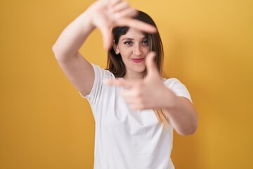 Poster - Young brunette woman standing over yellow background smiling making frame with hands and fingers with happy face. creativity and photography concept.