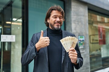 Sticker - Handsome middle age man holding 100 danish krone banknotes smiling happy and positive, thumb up doing excellent and approval sign