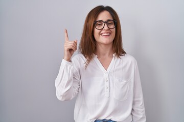 Sticker - Brunette woman standing over white isolated background pointing finger up with successful idea. exited and happy. number one.