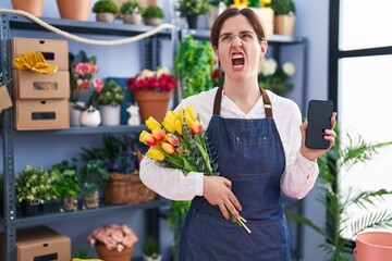 Sticker - Brunette woman working at florist shop holding smartphone angry and mad screaming frustrated and furious, shouting with anger. rage and aggressive concept.