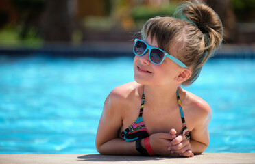 Young joyful child girl resting on swimming pool side with clear blue water on sunny summer day. Tropical vacations concept