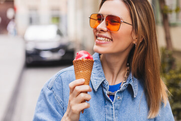 Close up happy young woman with delicious pink ice cream in waffle cone outdoors. Girl wear orange sunglasses in summer, spring or fall sunny day. Blonde female laughing and look at side.