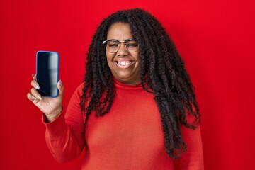 Poster - Plus size hispanic woman holding smartphone showing blank screen looking positive and happy standing and smiling with a confident smile showing teeth