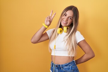 Poster - Young blonde woman standing over yellow background wearing headphones smiling looking to the camera showing fingers doing victory sign. number two.