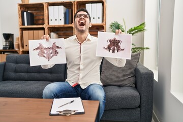 Sticker - Young hispanic man with beard holding rorschach test angry and mad screaming frustrated and furious, shouting with anger looking up.