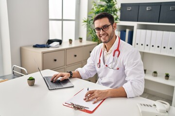 Wall Mural - Young hispanic man wearing doctor uniform using laptop working at clinic