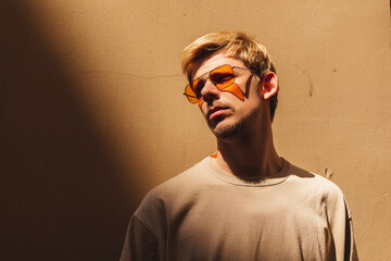 Handsome man standing near beige wall on the street of old city. Man look at side, wear orange sunglasses look at sun, beige t-shirt. Shadow and light of sunny summer, spring or fall day.