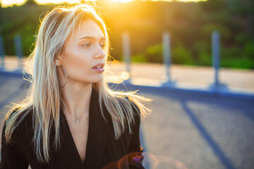 Close-up portrait of a beautiful young slim caucasian blonde girl in black dress dreamily standing in warm sunset light with softly blurred greenish background with copy space