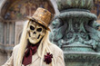 Venice, Veneto, Italy - Feb 19, 2023: Masquerade man in a costume of skull wearing a top hat and a suit during 2023 Venice Carnival celebrations. San Marco Basilica and fountain in the background