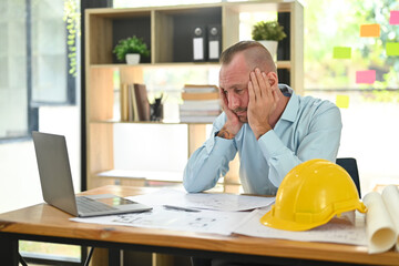 Stressed caucasian engineer man working with blueprints, examining construction plans at his workstation