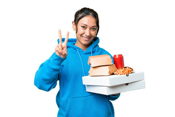 Wall Mural - Young Uruguayan woman holding pizzas and burgers over isolated chroma key background smiling and showing victory sign