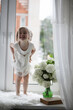 © alexkich - A little girl is sitting on the windowsill. A bouquet of flowers in a vase by the window and a girl sniffing flowers. A little princess in a white dress with a bouquet of white flowers by the window.