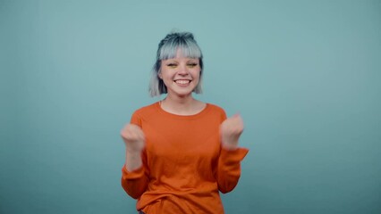 Wall Mural - Joyful hipster girl with blue hair stands on a blue background with a happy face raises her hands up and smiles, wears a green sweatshirt