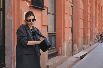 Wall Mural - Elegant young woman looking in her black leather bag her phone or purse. Business style woman wear grey blazer, black eyeglasses and bag on the street. Street style, fashion outfit.