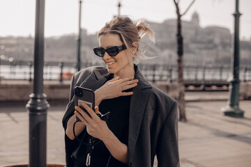 Wall Mural - Young blonde girl walk outdoors on the street and have a video call on her smartphone while smiling and point hand on herself, she look amazed, happy and shocking.