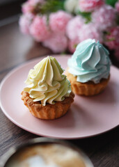 A Close-up of two Capcakes with Yellow and Blue Cream on a Pink Plate against a Wooden Table. A beige cup of cappuccino sits on the table. Near a plate of cakes lie beautiful pink and white flowers.