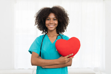 Portrait of smiling young female doctor or young nurse wearing blue scrubs uniform and stethoscope and standing holding red heart pillow while looking at camera isolated on white background