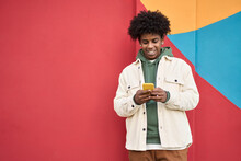 Happy Young African American Guy Standing At Color Bright Red Wall Outdoors Using Cell Phone, Looking At Camera Holding Cellphone Enjoying Doing Online Ecommerce Shopping In Mobile Apps, Playing Game.