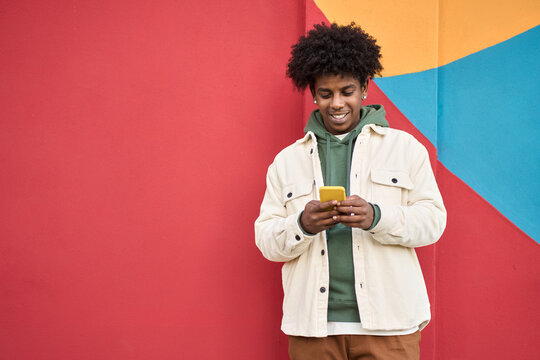 happy young african american guy standing at color bright red wall outdoors using cell phone, lookin