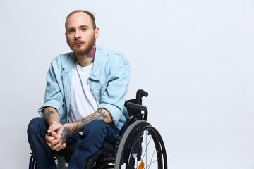 A man in a wheelchair looks thoughtfully at the camera, copy space, with tattoos on his arms sits on a gray studio background, the concept of health a person with disabilities, a real person