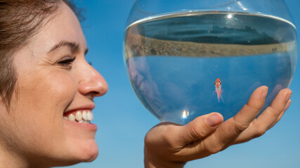 Wall Mural - Woman holding round aquarium with goldfish on blue sky background. 