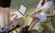 © Natee Meepian - Two beautiful Asian women in lovely dresses enjoying picnic in the park, sipping , work on laptop sitting on picnic chairs