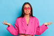 Indoor shot of hesitant clueless brunette woman shrug shoulders spread palms aside has doubt while make decision stands puzzled wears modern pink jacket and transparent spectacles isolated over blue
