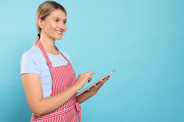 Sticker - Beautiful young woman in clean striped apron with tablet on light blue background. Space for text